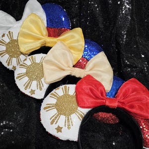 May include: Three pairs of Minnie Mouse ears with a white, red, and blue bow. Each pair features a white ear with a gold glitter sun and stars, representing the Philippine flag.