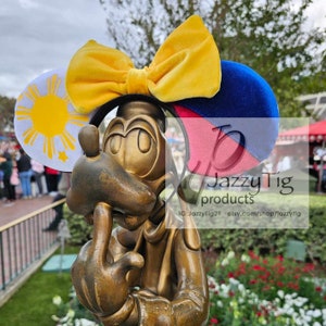 May include: A pair of Minnie Mouse ears with a yellow velvet bow, a white ear with a yellow sun and rays, and a blue and pink ear. The ears are on a bronze statue of a cartoon dog.