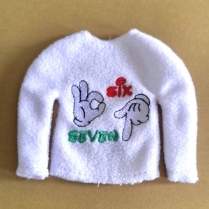May include: A white, long-sleeved shirt with the word "six" in red and "seven" in green. The shirt features embroidered hand gestures in black and white.