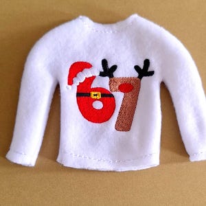 May include: A white felt Christmas sweater with long sleeves. The sweater features the number "67" in red and brown, with a Santa hat on the "6" and reindeer antlers above the numbers. A gold buckle accents the red number.