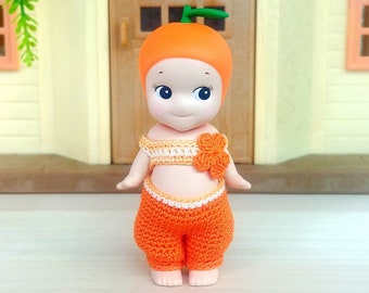 May include: A small orange doll with a crocheted orange and white outfit. The doll has a round orange head with a green stem on top and large blue eyes.