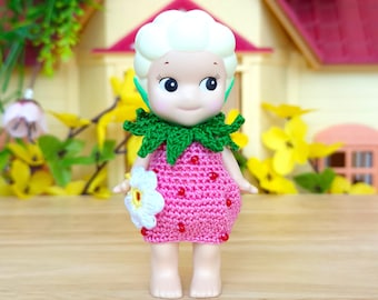 May include: A crocheted strawberry doll with a green leaf collar and a white flower in its hand. The doll has a pink and white crocheted dress and a white head with painted facial features.