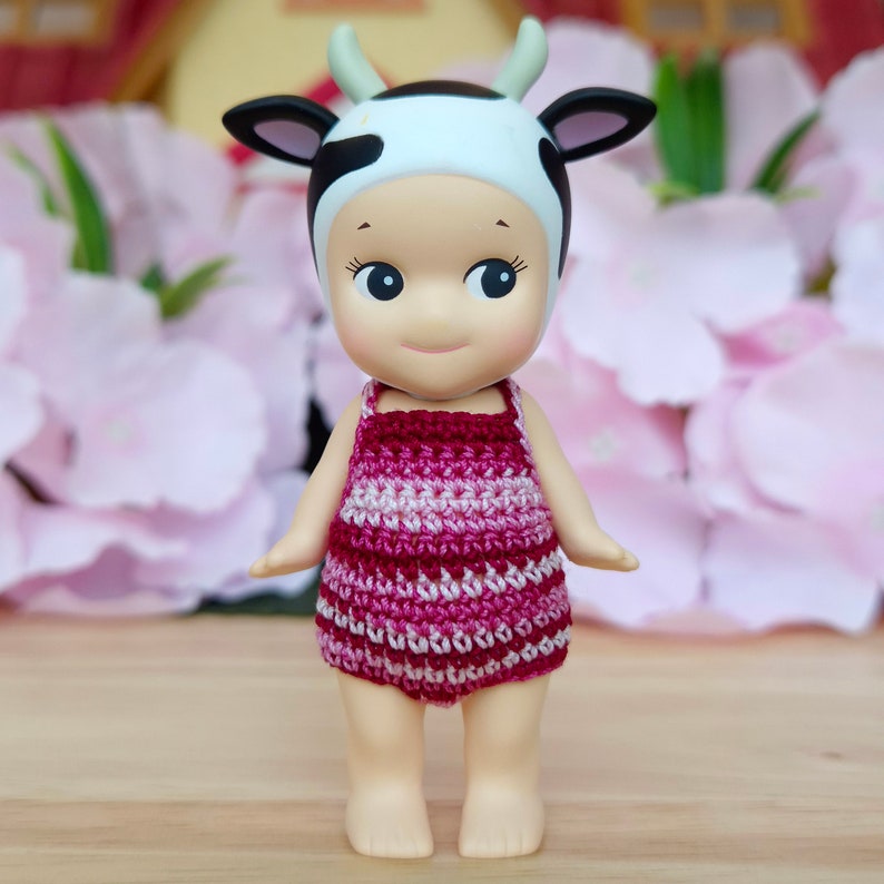 Puede incluir: Una peque&ntilde;a mu&ntilde;eca con un traje de ba&ntilde;o de crochet rosa y blanco y un sombrero de estampado de vaca. La mu&ntilde;eca tiene una cara sonriente y est&aacute; de pie sobre una superficie de madera.
