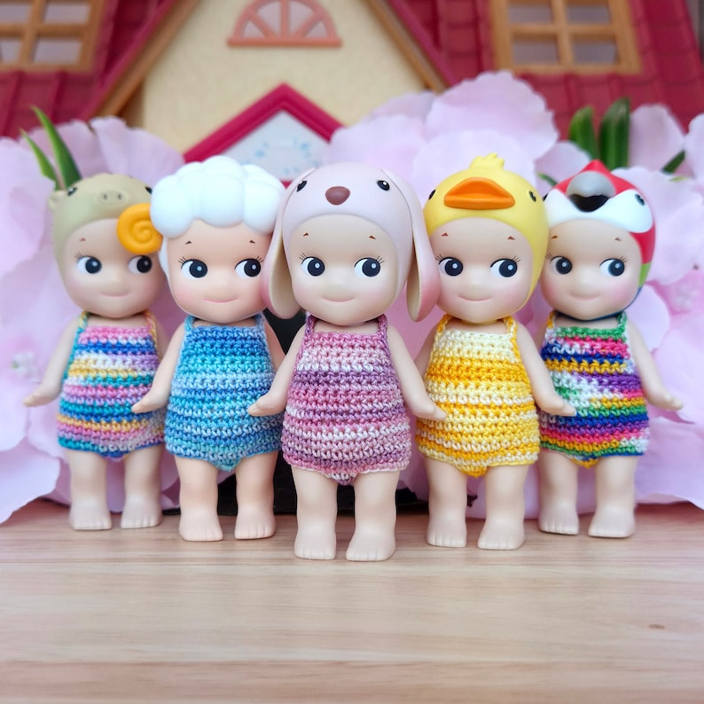 Puede incluir: Cinco peque&ntilde;as mu&ntilde;ecas con atuendos de crochet y sombreros con tem&aacute;tica animal. Las mu&ntilde;ecas est&aacute;n de pie sobre una superficie de madera. Las mu&ntilde;ecas llevan atuendos de crochet coloridos y sombreros con orejas de animales.