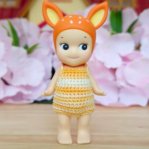 Puede incluir: Una peque&ntilde;a mu&ntilde;eca con cabeza de ciervo y un vestido de crochet amarillo y blanco. La mu&ntilde;eca tiene una cara sonriente y ojos grandes.