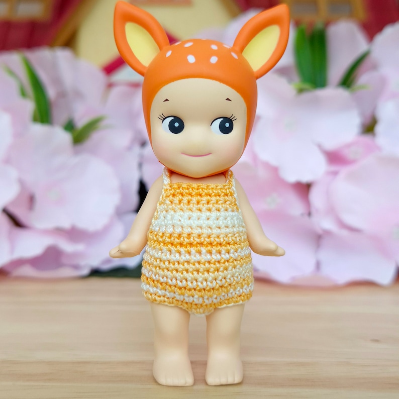 Puede incluir: Una peque&ntilde;a mu&ntilde;eca con cabeza de ciervo y un vestido de crochet amarillo y blanco. La mu&ntilde;eca tiene una cara sonriente y ojos grandes.