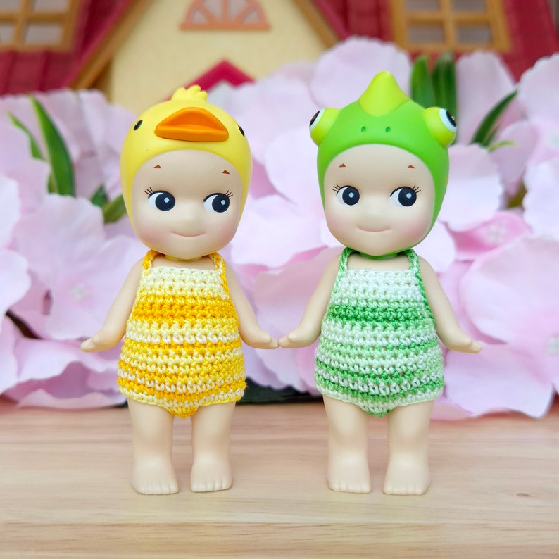 Puede incluir: Dos peque&ntilde;as mu&ntilde;ecas que llevan trajes de ba&ntilde;o de ganchillo y sombreros de animales. La mu&ntilde;eca de la izquierda lleva un sombrero de pato amarillo y un traje de ba&ntilde;o a rayas amarillo y blanco. La mu&ntilde;eca de la derecha lleva un sombrero de camale&oacute;n verde y un traje de ba&ntilde;o a rayas verde y blanco.