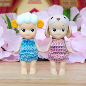 Puede incluir: Dos mu&ntilde;ecas peque&ntilde;as con atuendos de ganchillo. La mu&ntilde;eca de la izquierda lleva un atuendo a rayas azul y blanco y tiene una cabeza blanca y esponjosa con orejas amarillas. La mu&ntilde;eca de la derecha lleva un atuendo a rayas rosa y blanco y tiene un gorro de orejas de conejo marr&oacute;n.