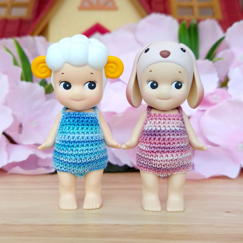 Puede incluir: Dos mu&ntilde;ecas peque&ntilde;as con atuendos de ganchillo. La mu&ntilde;eca de la izquierda lleva un atuendo a rayas azul y blanco y tiene una cabeza blanca y esponjosa con orejas amarillas. La mu&ntilde;eca de la derecha lleva un atuendo a rayas rosa y blanco y tiene un gorro de orejas de conejo marr&oacute;n.