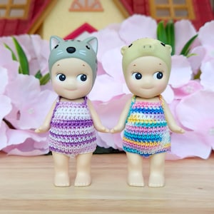 Puede incluir: Dos mu&ntilde;ecas peque&ntilde;as que llevan trajes de ba&ntilde;o de ganchillo. La mu&ntilde;eca de la izquierda lleva un traje de ba&ntilde;o de rayas blancas y moradas y un gorro de gato gris. La mu&ntilde;eca de la derecha lleva un traje de ba&ntilde;o de rayas multicolores y un gorro de cerdo verde.
