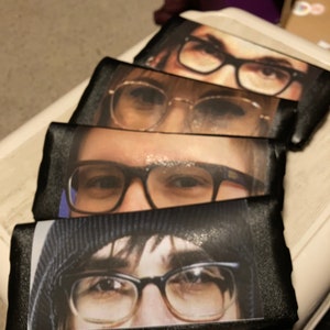 May include: Four black satin fabric bookmarks with different images of people wearing glasses. Each bookmark features a close-up of the person's eyes and glasses.