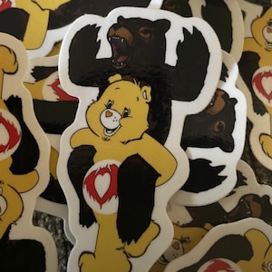 Puede incluir: Un oso de peluche amarillo con un corazón rojo en el pecho está siendo sostenido por un oso negro con las garras afuera. El oso de peluche sonríe y el oso negro gruñe. La imagen es una pegatina.