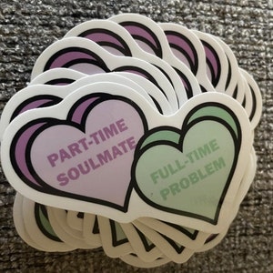 Könnte beinhalten: Ein Stapel herzförmiger Aufkleber mit dem Text "Part-time Soulmate, Full-time Problem" in einem hellvioletten und grünen Farbschema.