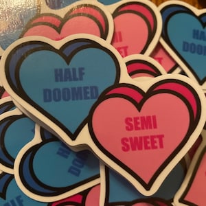 Puede incluir: Un conjunto de pegatinas en forma de corazón con diferentes lemas. Las pegatinas son azules y rosas con contornos negros. El texto en las pegatinas dice "Half Doomed" y "Semi Sweet".
