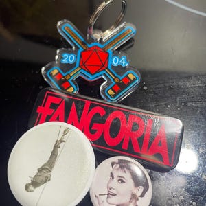 Könnte beinhalten: Eine Sammlung von vier Pins. Ein Pin ist ein roter und blauer Acryl-Schlüsselanhänger mit einem Würfel und gekreuzten Hockeyschlägern. Ein weiterer Pin ist ein rechteckiger Pin mit dem Wort "Fangoria" in weißen Buchstaben auf schwarzem und rotem Hintergrund. Die beiden anderen Pins sind rund und weiß mit einem Schwarzweißbild einer Person, die an einem Seil hängt, und einem Schwarzweißbild einer Frau mit einer Zigarette.