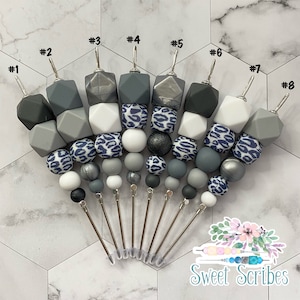 以下が含まれることがあります： グレー、白、ネイビーブルーの様々な色合いのビーズペン先8個セット。各ペン先は、幾何学的なビーズと丸いビーズの異なる組み合わせが特徴で、ヒョウ柄のデザインのものもあります。ペン先は扇形に配置されています。