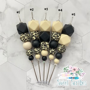 以下が含まれることがあります： 銀色の茎が付いた、ビーズ付きカクテルピック5本。ビーズは黒、クリーム色、ヒョウ柄です。上部のビーズは幾何学的な形をしています。ピックは白とグレーの模様の表面に配置されています。テキスト「Sweet Scribes」は右下にあります。