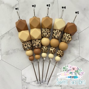 以下が含まれることがあります： 装飾ビーズのデザインが施された5つのカクテルピックのコレクション。各ピックは、茶色、タン、ヒョウ柄の色合いの幾何学的なビーズと丸いビーズのさまざまな組み合わせが施されたステンレス鋼の串が特徴です。