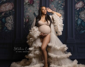 May include: A pregnant person wearing a white, ruffled gown with a floral backdrop. The gown has multiple layers of ruffles and is sheer. The person is standing with their hand on their stomach.