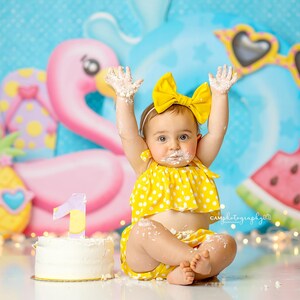 May include: A baby girl wearing a yellow polka dot dress and a yellow bow headband sits in front of a cake with a number one on top. The cake is decorated with white frosting. The background is a blue and pink tropical themed backdrop with a flamingo, watermelon, and pineapple.