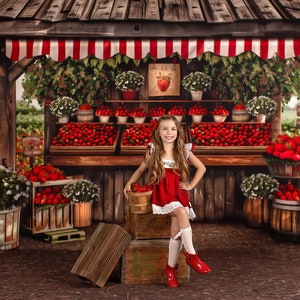 May include: A young girl in a red dress sits on a wooden crate in front of a strawberry stand. The stand is decorated with red and white striped awning and has shelves filled with strawberries. The girl is holding a basket of strawberries.