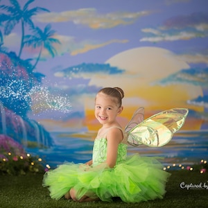 May include: A young girl dressed as a fairy with green wings and a green tutu sits in a grassy area in front of a painted backdrop of a tropical sunset with palm trees and a waterfall. The girl is smiling and looking at the camera. The text "Captured by Erin" is visible in the bottom right corner of the image.