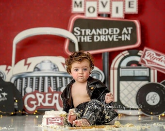 May include: A baby boy wearing a black leather jacket sits in front of a drive-in movie themed backdrop. He is holding a piece of cake with a red and white 'Rydell' logo. The backdrop features a white car, a jukebox, and a sign that reads 'Stranded at the Drive-In'.