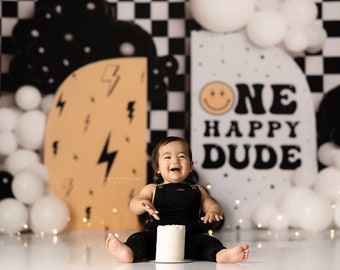 May include: A baby boy in black overalls sits in front of a black and white checkered backdrop with white balloons and a sign that says "One Happy Dude" with a smiley face.