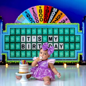 May include: A baby girl in a purple sequined dress and a purple bow sits in front of a Wheel of Fortune game board. The board says "It's My Birthday". The baby is eating a white cake with rainbow sprinkles.