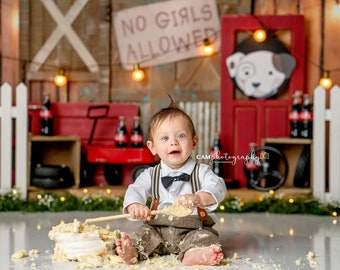 May include: A baby boy wearing a white shirt, brown pants, and a black bow tie sits on the floor in front of a cake. He is holding a wooden spoon and is covered in cake frosting. The background is a rustic wooden wall with a red wagon and a sign that says "No Girls Allowed".
