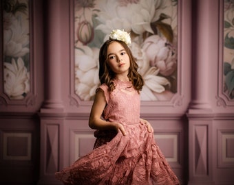 May include: A young girl wearing a pink lace dress with a white flower crown. The dress has a ruffled skirt and a high neckline. The girl is standing in front of a pink and white floral backdrop.