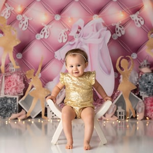 May include: A young girl wearing a gold sequined dress sits on a white stool in front of a pink and gold backdrop with a ballerina theme. The backdrop features a large pink ballet shoe, smaller pink ballet shoes, and gold ballerina silhouettes.