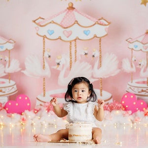 May include: A baby girl in a white dress sits in front of a pink carousel backdrop with white swans and pink hearts. She is holding a white cake with a pink heart on top.