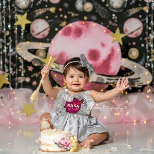May include: A baby girl wearing a silver sequined dress with a NASA patch and a silver bow in her hair is sitting in front of a space-themed backdrop. She is holding a wooden spoon and has cake on her face and hands.