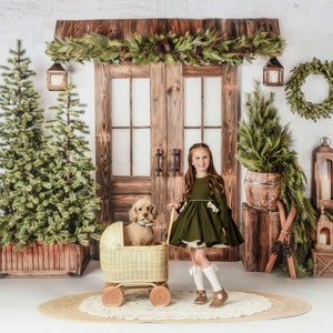 May include: A young girl in an olive green dress stands next to a vintage wicker doll carriage with a dog inside. The scene is set in front of a rustic wooden door decorated with greenery and Christmas trees, creating a festive holiday atmosphere.