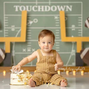 May include: A baby in a brown and white striped jumpsuit sits in front of a green and yellow football field backdrop. The baby is covered in cake frosting and is holding a piece of cake in their hand. The backdrop has the word "TOUCHDOWN" written on it.