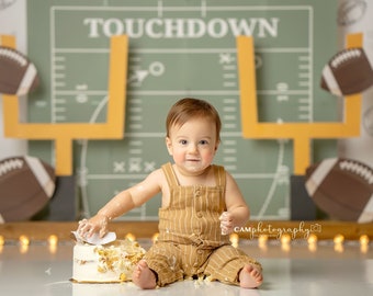 May include: A baby in a brown and white striped jumpsuit sits in front of a green and yellow football field backdrop. The baby is covered in cake frosting and is holding a piece of cake in their hand. The backdrop has the word "TOUCHDOWN" written on it.