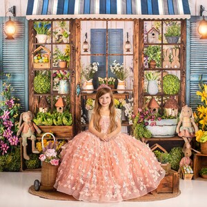 May include: A young girl wearing a pink princess dress sits in front of a floral backdrop. The backdrop features a wooden storefront with a blue awning and a window with blue shutters. The girl is smiling and has long blonde hair.