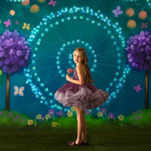 May include: A young girl in a purple and grey tulle dress holds a red lollipop in front of a whimsical backdrop featuring a blue ferris wheel, purple trees, and green foliage.