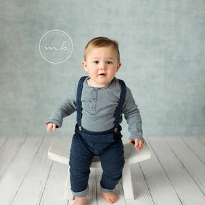 May include: A baby boy wearing a blue and white striped shirt with suspenders and blue pants sits on a white stool. The background is a light blue textured wall.