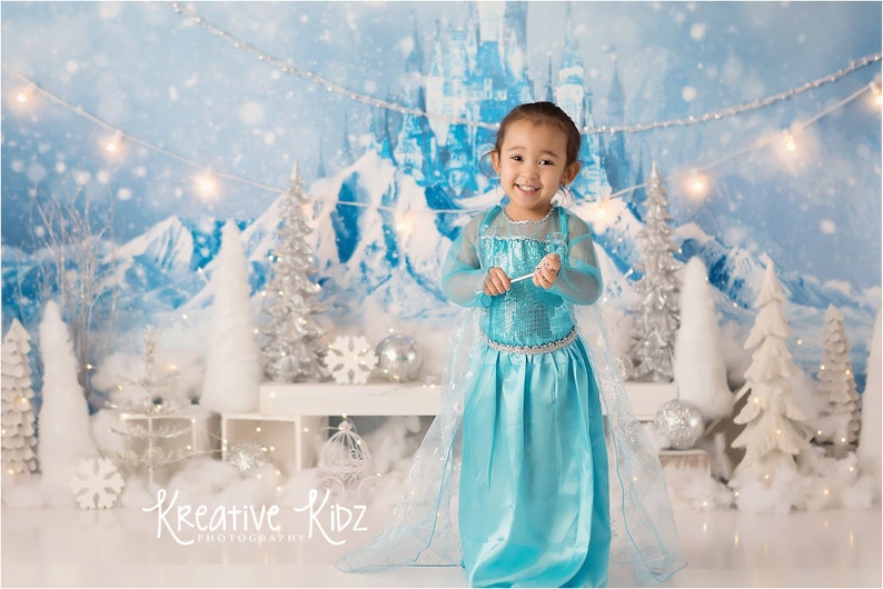 Pu&ograve; includere: Una bambina vestita da principessa con un abito blu, una cintura argentata e una bacchetta argentata, in piedi davanti a uno sfondo invernale con un castello sullo sfondo. Lo sfondo presenta alberi bianchi, neve e una ghirlanda di luci.