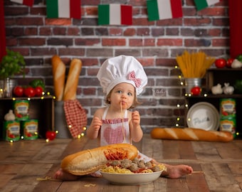 May include: A young child dressed as a chef is eating spaghetti with meatballs. The child is wearing a white chef hat with a red bow and a red and white checkered bib. The child is sitting on a wooden floor in front of a brick wall with Italian flags hanging above. There is a baguette and a bowl of spaghetti with meatballs in front of the child.