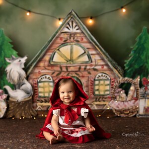 May include: A young girl dressed as Little Red Riding Hood sits in front of a whimsical backdrop featuring a gingerbread house, a wolf, and a basket. The backdrop is decorated with string lights.