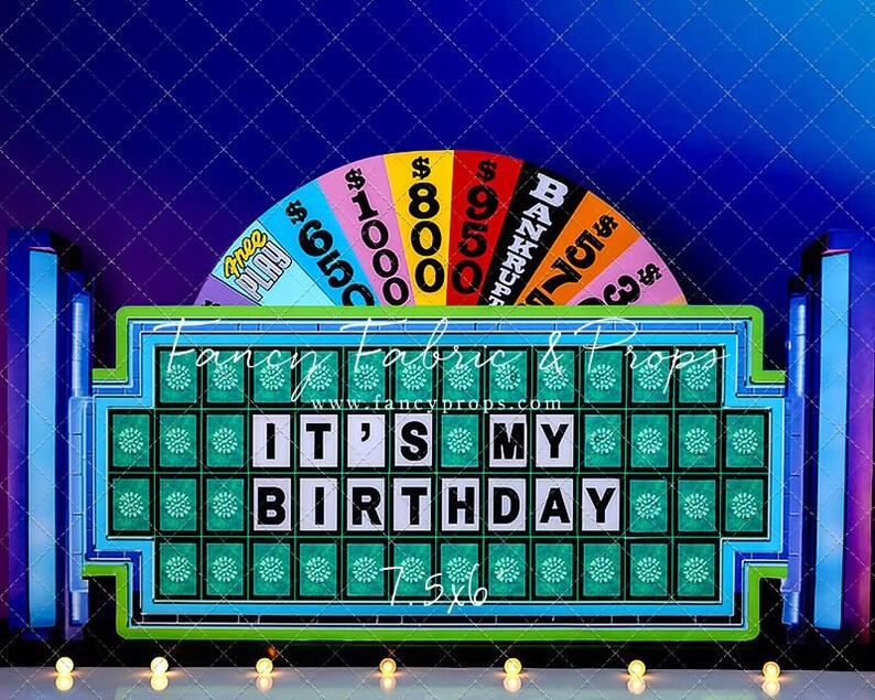 May include: A blue and green cardboard cutout of a Wheel of Fortune game board with the phrase "It's My Birthday" in white letters. The board has a green background with a grid of squares and a spinning wheel with dollar amounts and the word "Bankrupt".