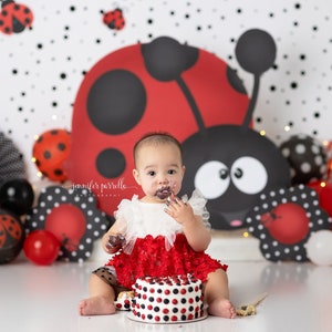 May include: A baby girl in a red and white dress is sitting in front of a ladybug-themed backdrop and eating a red and white polka dot cake. The baby has frosting on her face and hands.