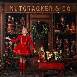 May include: A young girl in a red dress stands in front of a toy shop called "Nutcracker & Co." The shop is decorated with Christmas lights and greenery. The girl is holding a red wagon filled with nutcracker figurines. Snow is falling in the background.