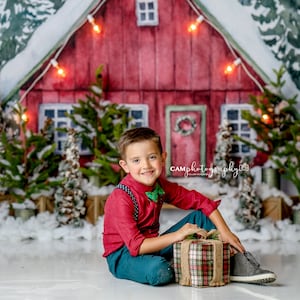 May include: A young boy in a red shirt and blue pants sits in front of a red barn with a snowy backdrop. He is holding a wrapped gift with a plaid pattern and a brown bow. The barn has a wreath on the door and lights strung across the front.