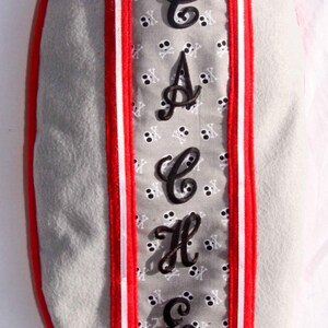 May include: A gray fabric bag with red and white stripes and a black embroidered name "CACHE" on the front. The bag has a small black loop at the top and a white drawstring.