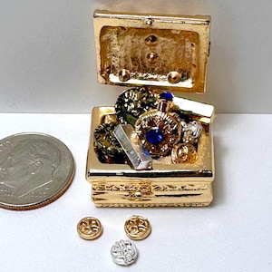 Könnte beinhalten: Eine kleine goldene Schatztruhe mit einem Scharnierdeckel ist geöffnet und zeigt eine Vielzahl von kleinen Schmuckstücken und Juwelen im Inneren. Die Truhe steht auf einer weißen Oberfläche, auf der drei goldene und silberne Münzen verstreut liegen.