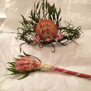 May include: A mermaid crown and wand made with seashells, greenery, and pearls. The crown is a wire base with a large seashell in the center, surrounded by greenery and pearls. The wand is a wooden stick wrapped in twine and decorated with a seashell, greenery, and pearls.